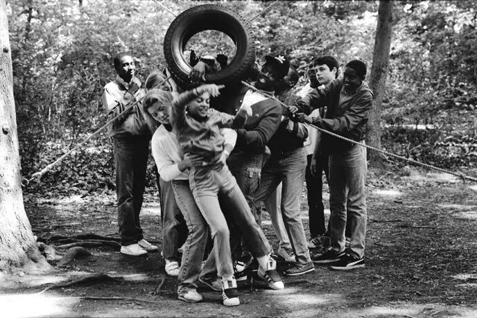 The image shows a group of people in a forest setting. They appear to be engaged in a team-building activity, possibly a game. One person is holding a tire above their head, while others are interacting with them. The photo is in black and white, suggesting an older time.